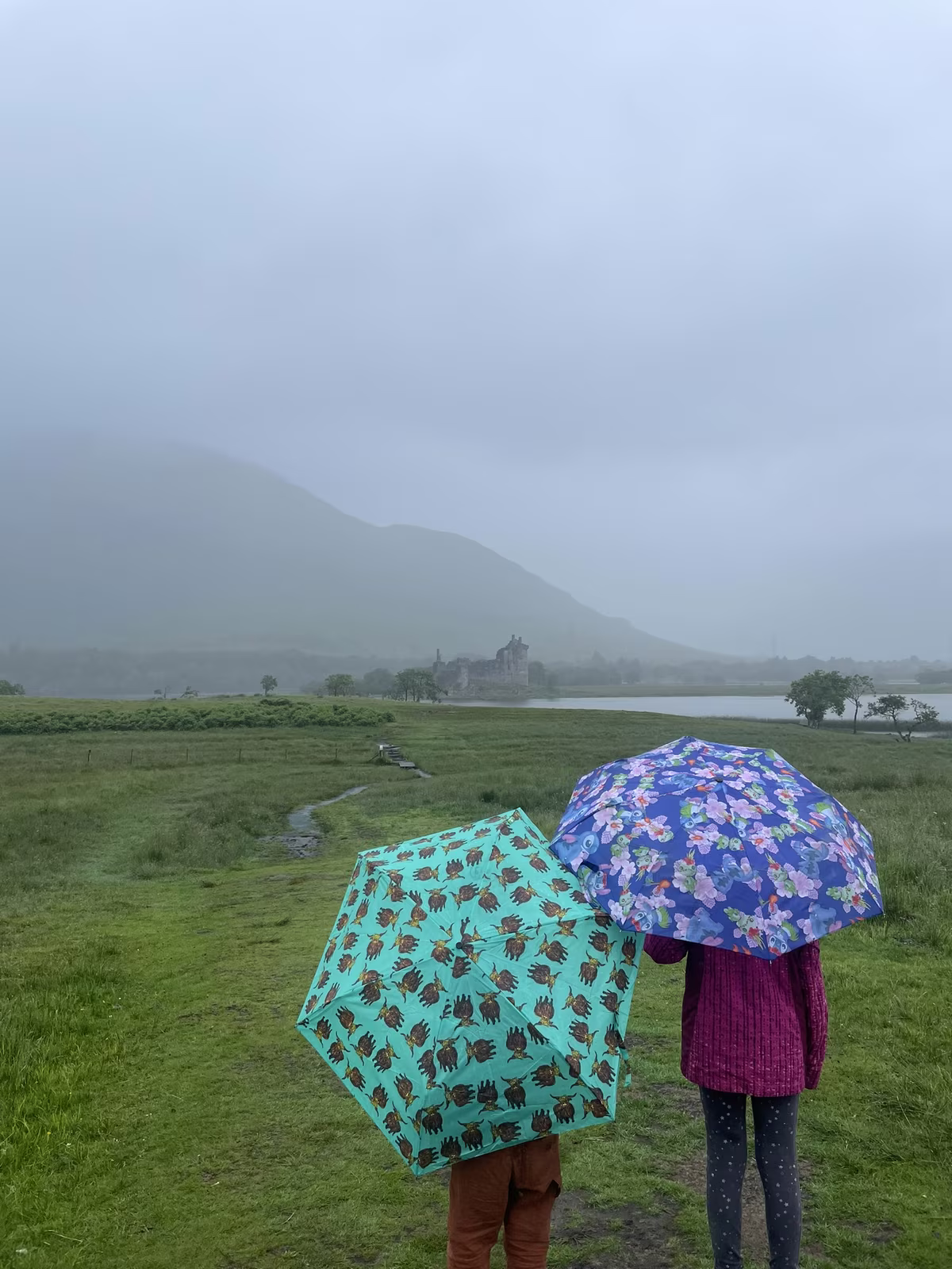 Deux enfants sous des parapluies colorés face au château de Kilchurn sous la pluie